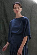 Load image into Gallery viewer, A woman with dark hair in a low bun models the Kimono Top | Sandwashed Silk Charmeuse Teal, featuring wide sleeves, as she stands before a textured dark green backdrop and looks calmly at the camera.