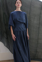 Load image into Gallery viewer, A woman stands indoors, calmly looking ahead while wearing the Hand Pleated Column Wrap Skirt in sandwashed silk charmeuse teal; wide sleeves and a cinched waist complete her look, set against a sheer textured dark backdrop.