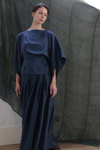 Load image into Gallery viewer, A woman wearing the Hand Pleated Column Wrap Skirt in sandwashed silk charmeuse teal stands before a sheer, dark green textured backdrop. She has short brown hair and gazes slightly downward with a neutral expression.