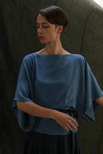 Load image into Gallery viewer, A woman with short dark hair stands against a dark, textured background wearing the Kimono Top | 2-ply Silk Crepe Ai Dye in blue, its rich hue suggesting an indigo-dyeing process. She looks down calmly in the loose-fitting style.