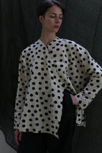 Load image into Gallery viewer, A person with short hair wears the Qipao Blouse | Polkadot Seersucker Ivory and black pants, standing against a dark draped background. Their hand rests on their hip as they look slightly downward.