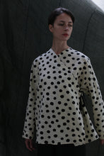 Load image into Gallery viewer, A woman with short dark hair wears the Qipao Blouse | Polkadot Seersucker Ivory, standing before a dark fabric backdrop. She looks down calmly, highlighting the blouse’s cream color and black polka dot pattern.
