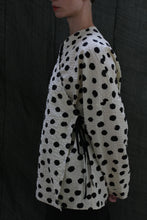 Load image into Gallery viewer, A person stands sideways wearing the Qipao Blouse | Polkadot Seersucker Ivory—a cream wrap top with black polkadots and side ribbon ties—against a dark, textured fabric background.