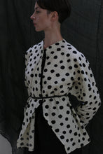 Load image into Gallery viewer, A person with short dark hair models the Qipao Blouse | Polkadot Seersucker Ivory, featuring large black polkadots and a thin black waist belt, standing in profile before a dark, draped backdrop.