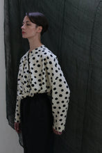 Load image into Gallery viewer, A person with short hair wears the Qipao Blouse | Polkadot Seersucker Ivory, styled with high-waisted dark pants, standing before a draped dark fabric background.