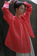 Load image into Gallery viewer, A person with short dark hair stands with arms raised, wearing the Qipao Blouse | Silk Organza Sakura in bright pink and light-colored pants, set against a dark, textured background.