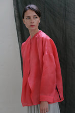 Load image into Gallery viewer, A person with short dark hair wears the Qipao Blouse | Silk Organza Sakura, a loose, bright pink long-sleeved top, standing before a dark textured background and gazing slightly to the side with a neutral expression.