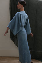 Load image into Gallery viewer, A person with short dark hair poses in profile indoors, wearing the Kimono Dress | Silk Noil Ai Dye—an ankle-length, loose blue dress with wide sleeves and a kimono-inspired design—against a dark curtain backdrop.