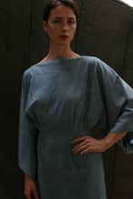 Load image into Gallery viewer, Someone poses with one hand on their hip against a dark, textured background, wearing the Kimono Dress | Silk Noil Ai Dye, a loose-fitting dress in light blue with a subtle kimono-inspired design.