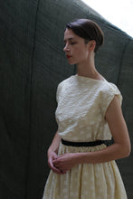 Load image into Gallery viewer, A woman with short brown hair wears the Magnolia Dress | Polkadot Seersucker Butter, a cream-colored dress with a subtle polkadot pattern and black belt, standing before a dark green fabric backdrop, looking down with hands gently clasped.