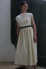 Load image into Gallery viewer, A woman stands indoors against a dark fabric backdrop, looking slightly to the side, wearing the Magnolia Dress | Polkadot Seersucker Butter—an ankle-length, light polkadot dress with a black belt.