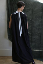 Load image into Gallery viewer, A person with short dark hair stands facing away, wearing the Shoulder Pleat Dress in Clip Dot Black—a long, sleeveless black gown with a white neck ribbon. The textured black clip dot fabric hangs in the background against a white wall and wood floor.