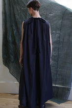 Load image into Gallery viewer, A person with short brown hair stands with their back to the camera, wearing the Shoulder Pleat Dress in Clip Dot Black; a textured dark green cloth hangs behind them.