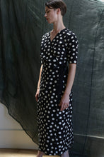 Load image into Gallery viewer, A person stands indoors against a dark, textured fabric backdrop, wearing the Fitted Wrap Dress | Polkadot Seersucker Black, featuring white polka dots, short sleeves, and a side tie, looking slightly downward with hands relaxed by their sides.