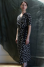 Load image into Gallery viewer, A woman with short dark hair and a neutral expression stands indoors wearing the Fitted Wrap Dress | Polkadot Seersucker Black, a black calf-length dress with white polka dots and a sash belt, set against a backdrop of dark textured fabric.