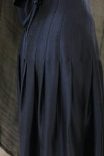 Load image into Gallery viewer, Close-up of the Hand Pleated Column Wrap Skirt in sandwashed silk charmeuse teal, featuring vertical stitched lines and pleats. The background is blurred with a softly textured, neutral tone.