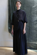 Load image into Gallery viewer, A person wears the Hand Pleated Column Wrap Skirt in Silk Charmeuse Black, paired with a loose black button-up top, standing indoors against a dark green fabric backdrop with soft natural light and wooden floors.