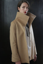 Load image into Gallery viewer, A person with short dark hair stands in front of a dark background, wearing the Stamford II | Camel over a white shirt. The signature jacket, crafted from 100% woven cashmere, drapes perfectly as the individual's head is slightly tilted, exuding an air of confidence and poise.