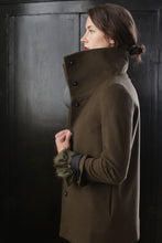 Load image into Gallery viewer, A woman in an olive green cashmere coat stands in profile with the collar turned up. She has her dark hair tied in a low bun and holds the coat with her left hand. The background is dark and features a textured surface, highlighting the elegance of this Stamford II | Olive jacket.