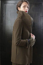 Load image into Gallery viewer, A person with dark hair styled in an updo, wearing a high-collared Stamford II | Olive coat, stands against a dark background. They glance over their shoulder with a serious expression. The coat's cuff features a fur-like embellishment.