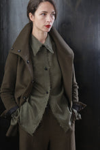 Load image into Gallery viewer, A person wearing a stylish, dark olive green Stamford II | Olive jacket over a matching top and pants stands with hands in their pockets. They have short, dark hair, a neutral expression, and are positioned against a dark, glossy background.