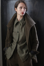 Load image into Gallery viewer, A person with short, dark hair stands against a dark background, dressed in a Stamford II | Olive jacket with a stylish high collar and a gray shirt underneath. They have their hands in their pockets and wear a serious expression.