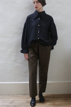 Load image into Gallery viewer, A person stands against a plain light-colored wall, wearing a loose black button-up shirt with frayed cuffs and collar, Cashmere Unisex Pants in Olive with a silk ribbon drawstring, and black shoes. They have short dark hair styled neatly, red lipstick, and their left hand in their pocket.