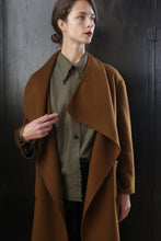 Load image into Gallery viewer, Woman with short brown hair wearing a long, tobacco-colored coat over a green collared shirt. The Winston Long Double-face showcases relaxed construction as she stands against a dark, textured background with her left hand holding the lapel. She looks slightly to the right with a neutral expression.