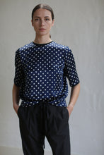 Load image into Gallery viewer, A person with straight, pulled-back hair stands against a plain background. They are wearing a Crewneck Tee | Navy Polkadot and black pants. Their hands rest in their pants pockets, adding to the casual elegance of their look.