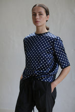Load image into Gallery viewer, A person with their hair tied back is looking at the camera while wearing a Crewneck Tee | Navy Polkadot and black pants with hands in pockets. The background is plain and softly lit.