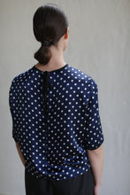 Load image into Gallery viewer, A person with dark hair tied in a low bun is shown from the back. They are wearing a Crewneck Tee | Navy Polkadot. The background is plain and neutral, focusing attention on the person and the pattern of their blouse.