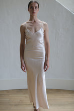 Load image into Gallery viewer, A person stands indoors in front of a white wall and a wooden floor. They are wearing the Liquid Slip Dress Long | Champagne, made from biodegradable plant-derived material, with thin straps. Their expression is neutral, and they have short, dark hair pulled back.