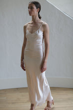 Load image into Gallery viewer, A person with tied-back dark hair wearing a Liquid Slip Dress Long | Champagne stands on a wooden floor against a light-colored wall. They are looking to the side and have a poised and calm demeanor.