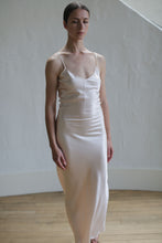 Load image into Gallery viewer, A woman stands in a minimalist indoor setting, wearing the Liquid Slip Dress Long | Champagne made from biodegradable, plant-derived material with thin spaghetti straps. Her hair is slicked back, and she has a calm, serene expression. The background features a plain, textured white wall and light-colored wooden flooring.