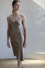Load image into Gallery viewer, A woman stands against a light-colored, textured wall, wearing a Liquid Slip Dress | Mink with thin straps. She has long dark hair pulled back and gazes slightly to the left. Sunlight softly illuminates the scene from an unseen source, highlighting the eco-friendly allure of her plant-derived material outfit.