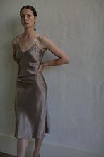 Load image into Gallery viewer, A person with long dark hair, pulled back, is standing against a textured, neutral-colored wall. They are wearing the Liquid Slip Dress | Mink made from plant-derived material, its silky bronze color and spaghetti straps adding to the allure. One hand rests on their hip while the other hangs by their side.