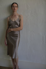 Load image into Gallery viewer, A woman with dark hair tied back is standing against a textured white wall, wearing a simple, elegant Liquid Slip Dress | Mink made of plant-derived material that reaches below her knees. Her gaze is directed toward the camera, and one hand rests on her waist while the other hangs by her side.