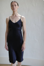 Load image into Gallery viewer, A person with long hair is wearing a Liquid Slip Dress | Midnight made from biodegradable material and standing against a plain, light-colored background. The elegant dress features thin shoulder straps and a V-neckline. The individual has a calm and poised expression.