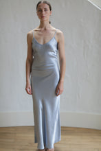 Load image into Gallery viewer, A woman stands against a plain background wearing a Liquid Slip Dress Long | Aluminum. She looks forward with a neutral expression. The floor beneath her is wooden, and the wall behind her is a light color with visible texture.