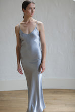 Load image into Gallery viewer, A person with straight, pulled-back hair is wearing a Liquid Slip Dress Long | Aluminum made from biodegradable material. They are standing on a wooden floor in front of a white, textured wall, looking slightly to the side. The dress is long and form-fitting with thin straps.