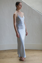 Load image into Gallery viewer, A woman stands on a wooden floor, wearing a long, form-fitting Liquid Slip Dress Long | Aluminum with thin straps. This sleek, sexy dress drapes elegantly around her figure. Her hair is pulled back, and she gazes forward with a neutral expression. The background features an off-white, textured wall that curves slightly upward.