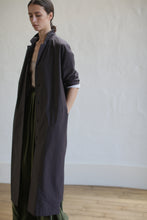 Load image into Gallery viewer, A person stands indoors wearing a brown Painter's Coat over a green skirt, accented by a leather Obi belt. They have one hand in their pocket and gaze downward. The background is a plain, light-colored wall and wooden floor.