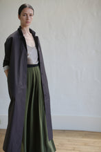 Load image into Gallery viewer, A person stands confidently in a stylish long brown "Painter's Coat," cinched at the waist with a sleek leather Obi belt, paired with a shimmering top and flowing green skirt in a minimalistic room with wooden flooring and plain walls.