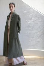 Load image into Gallery viewer, A person stands indoors by a textured white wall, wearing the Painter's Coat in Olive over a beige top and cream wide-legged pants. With brown hair pulled back, they pose in natural light, sandals on their feet.