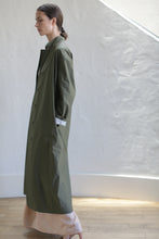 Load image into Gallery viewer, A woman with dark hair tied back wears the Painter's Coat | Olive over a light dress, standing in profile indoors against a textured white wall and wooden floor.