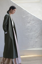 Load image into Gallery viewer, A woman with dark hair tied back, wearing the Painter's Coat | Olive over a light pink dress, stands indoors by a textured white wall as diagonal sunlight casts gentle shadows.