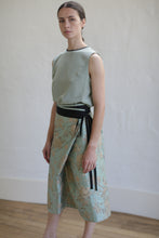Load image into Gallery viewer, A person wearing a sleeveless, light green top tucked into the limited edition Dupioni Petal Wrap Skirt in Jade stands against a plain wall. The outfit showcases pastel colors and an elegant, zero-waste design. The wooden floor contrasts with the neutral background.
