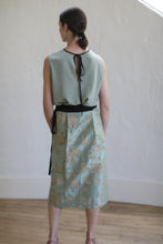 Load image into Gallery viewer, A person in a sleeveless light green top made of Dupioni silk with a bow at the back, paired with the limited edition Dupioni Petal Wrap Skirt in Jade. They face away on a wooden floor beside a textured white wall, highlighting their exquisite style.