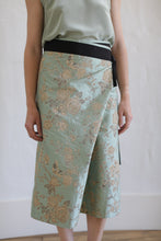 Load image into Gallery viewer, The person is wearing the Dupioni Petal Wrap Skirt in jade, featuring a floral pattern and a black waistband, crafted from textured Dupioni silk. They pair it with a matching sleeveless top. The photo showcases this limited edition ensemble indoors against a white background.