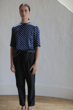 Load image into Gallery viewer, A person stands indoors against a plain, light-colored wall. They are wearing a navy blue shirt with white polka dots and Typewriter Unisex Pants | Black. The person has a neutral expression and their hair is pulled back with a Japanese grosgrain ribbon. The floor appears to be wooden.